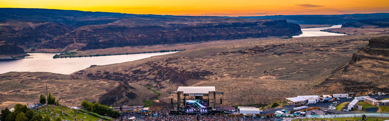 Panoramic view of The Gorge Amphitheatre in Washington at sunset, showing the stage, river, cliffs, and vast canyon landscape.