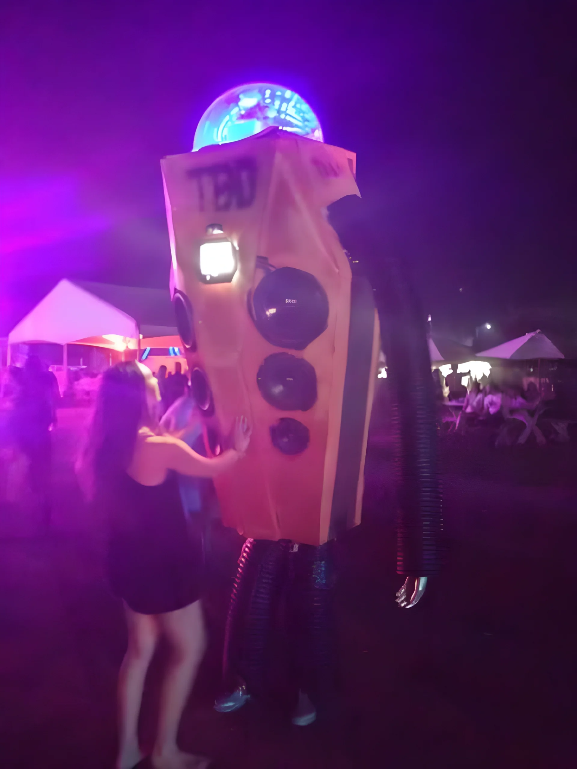 A festival attendee's homemade cardboard and foam recreation of the TBD Fest mascot Tibby the robot, worn at the 2015 festival, with glowing disco-ball head and illuminated speaker chest, based on Jason Malmberg's original character design<br />
